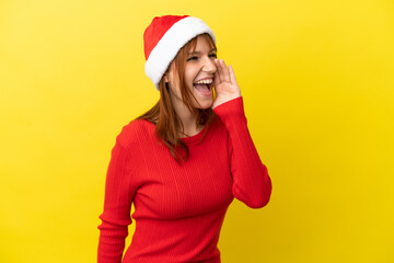 Redhead girl with christmas hat isolated on yellow background shouting with mouth wide open to the side