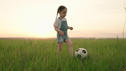 little kid plays football at sunset, baby runs around green football field and kicks ball, child...