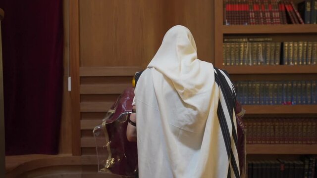 A Jew prays in the synagogue. A man stands near the presidium and reads a book. Rear view