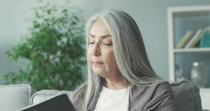 Close up of beautiful gray-haired woman sitting alone at home on couch and browsing new magazine looking at interesting photos and headlines.