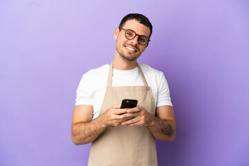 Brazilian restaurant waiter over isolated purple background sending a message with the mobile