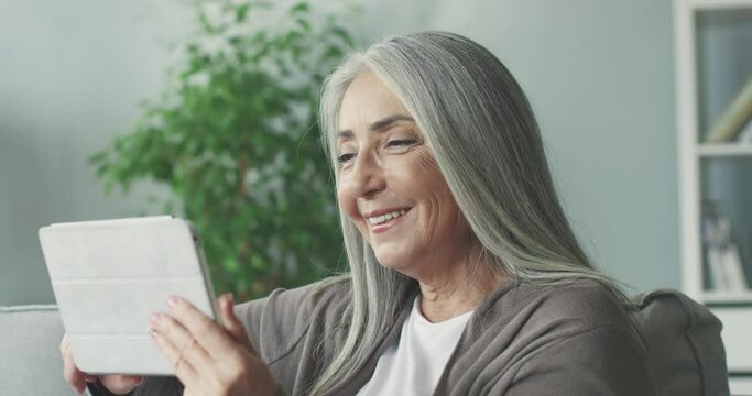 Close up of beautiful gray-haired woman sitting alone at home on couch and browsing online photos of her grandchildren smiling holding tablet.
