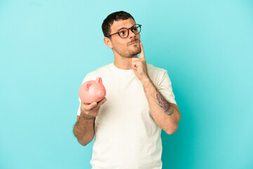 Brazilian man holding a piggybank over isolated blue background having doubts while looking up