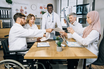 Fototapeta premium African american doctor in lab coat smiling on camera while his mixed race colleagues examining reports, graphs and x ray scan. Concept of cooperation and medicine.