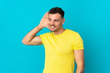 Young caucasian handsome man isolated on blue background listening to something by putting hand on the ear