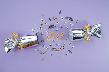Banner. Happy New Year and Merry Christmas. A silver foil firecracker with a gold number 2022 and confetti on a fashionable purple background. Flat lay.