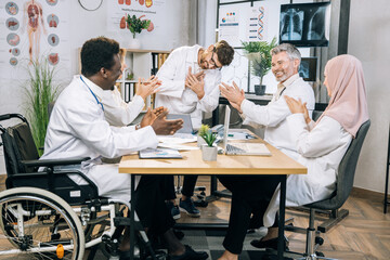 Team of multi ethnic doctors clapping to male colleagues after speech during conference. Happy medical specialists with one in wheelchair sitting at boardroom and coworking.