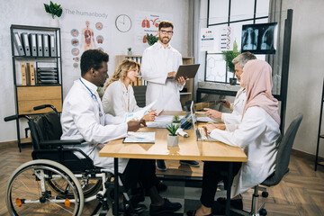 Caucasian doctor standing at boardroom with laptop in hands and looking at camera. Group of multicultural colleagues sitting at table with notes and gadgets. Concept of international conference.