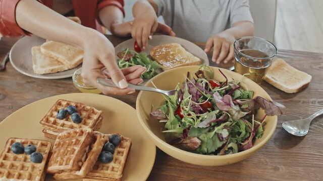 High-angle medium close-up with slowmo of unrecognizable woman taking care of her little son putting him fresh vegetables on plate while having breakfast together at home