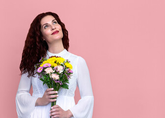 Woman Holding Flowers Looking Up with Soft Smile on Pink Background. Close-up portrait of beautiful happy woman in white dress holding bouquet of flowers. Room for copy.