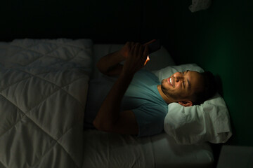 Cheerful young man texting in bed