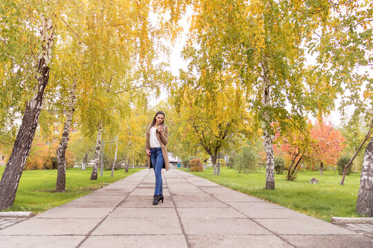 Young modern woman in a beige polto walks in an autumn park with coffee in her hands. Full-length photo. autumn walk with takeaway coffee. Fall park