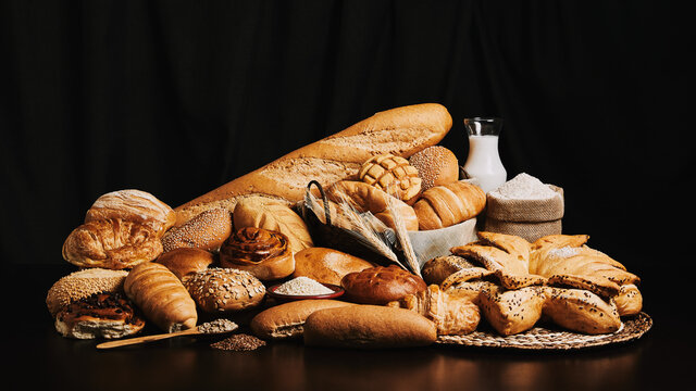 Variety of delicious breads and buns with ears of wheat, milk and flour on black background. Food and bakery concept with copy space.
