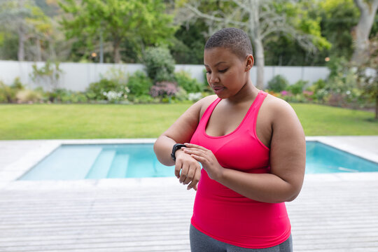 African american plus size woman in sports clothes looking at smartwatch in garden