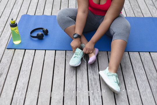 Midsection of african american plus size woman in sports clothes sitting on yoga mat and tying shoes