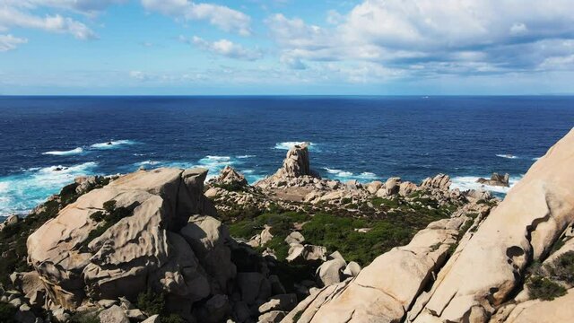 Aerial view forward, flying trouth two big rocks on the coastline, behind a noher big rock, the waves break into the bay. shaped by the water of the mediterranean sea and the wind.