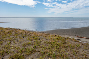 intersection of grass covered ground and sandy beach by the coast with clouds above the horizon over the sea