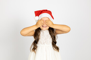 portrait of joyful girl with long hair in white dress, in red Santa Claus hat covering eyes with palms, isolated on white background, concept of waiting for New Year gift. 