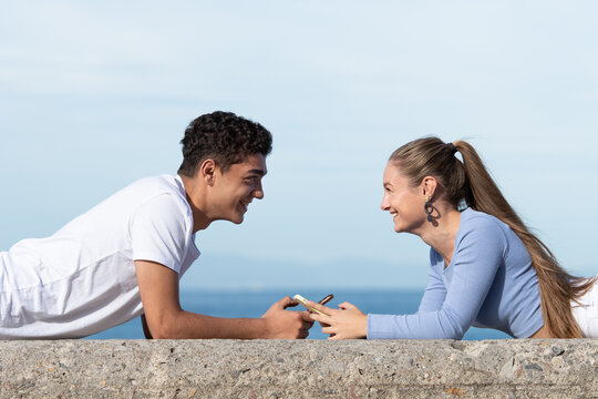 Teenager multiethnic couple looking each other and lying face to face while holding their phones.