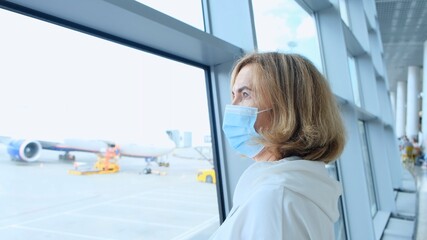 adult Female wearing protective mask is standing at the window in the airport terminal awaiting the departure of a flight due to travel restrictions due to the coronavirus pandemic, a senior aged 50