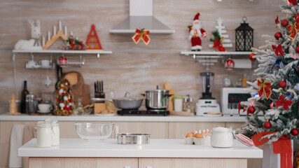 Empty christmas decorated culinary kitchen with nobody in it ready for traditional festive holiday. On table standing homemade cookie dessert ingredients with glass of milk, strainer