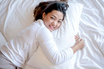 A young Asian woman is comfortable in bed, hugging a soft pillow in a hotel room with a bright, smiling, happy face. It is a good rest on a long vacation. Top view