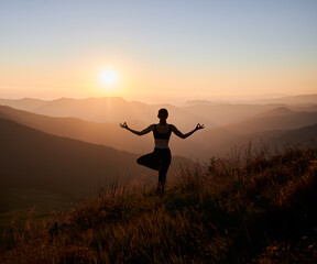 Back view of slim woman performing yoga pose on grassy hill with orange sky on background. Fit...