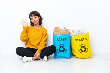 Young girl holding a bag full of plastic and paper sitting on the floor isolated on white background counting five with fingers