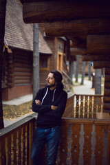 man with a beard in a black jacket and blue jeans stands at a wooden house in the forest