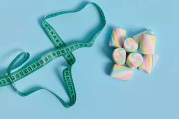 Top view of sweet colorful marshmallows and measuring tape on blue background with copy space for medical advertising. Link between overweight and unhealthy eating habits. World diabetes day concept