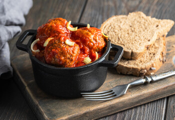 Meatballs with tomato sauce in the pot on wooden cut board with bread and fork.