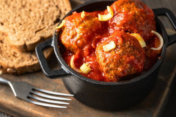 Close up of beef meatballs with tomato sauce in the pot on wooden cut board with bread and fork.