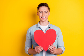 Portrait of attractive cheerful guy holding in hands big heart card isolated over bright yellow color background