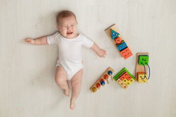 crying newborn baby in white bodysuit lies on his back on the floor and plays with educational toys. products for children. concept of a happy childhood and motherhood. child care. space for text
