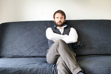 A bearded photographer is sitting on a couch and posing with his digital camera. He is wearing a grey vest and a white shirt.