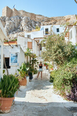 Fototapeta na wymiar Narrow greek street with many plants and houses on the mountain and a girl in a hat. Passion for travel and tourism. Back view of the girl.