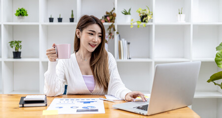 Beautiful Asian businesswoman sitting in her private office, drinking coffee and looking at information on her laptop, she is the female executive of a startup company. Concept of financial management