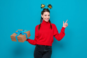 Happy caucasian smiling woman in red sweater celebrate new year party holds gift and point of finger on blue isolated background.