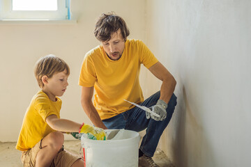 Man with his son makes repairs at home, he teaches children to plaster the walls with a spatula in...