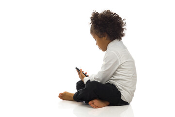 Little boy in elegant clothes is sitting on a floor and looking at telephone. Side view studio shot. - Powered by Adobe