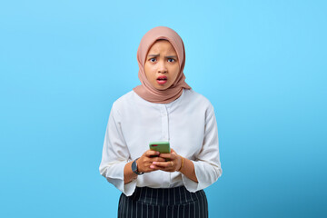 Portrait of shocked young Asian woman with open mouth using mobile phone over blue background