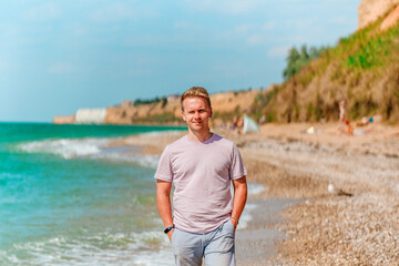 A young blond man walks on the beach near the azure ocean or sea. The concept of a happy summer vacation