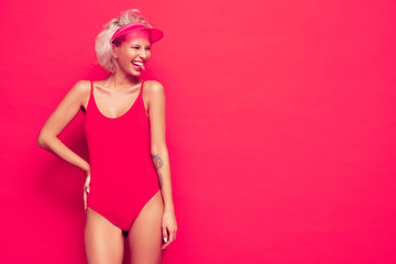 Portrait of young smiling blond model in summer swimwear bathing suit and transparent visor cap. Sexy carefree woman having fun and going crazy. Female posing near pink wall in studio. Shows tongue