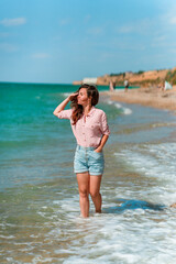 Fototapeta na wymiar A charming young woman with long hair and wearing a shirt walks along the beach on a sunny day along the azure sea or ocean. The concept of summer holidays