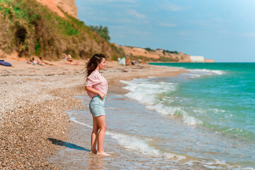 A charming young woman with long hair and wearing a shirt walks along the beach on a sunny day along the azure sea or ocean. The concept of summer holidays