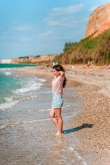 A charming young woman with long hair and wearing a shirt walks along the beach on a sunny day along the azure sea or ocean. The concept of summer holidays