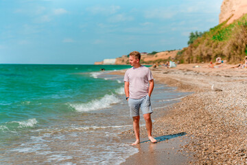 A young blond man walks on the beach near the azure ocean or sea. The concept of a happy summer vacation