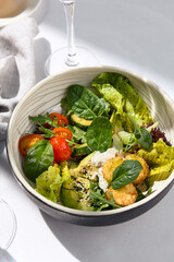 Salad bowl with avocado, tomato and cheese balls. Healthy vegetarian lunch with water glass on white table. Summer day dinner, sunlight and harsh shadow. Healthy salad bowl food. Organic salad bowl.