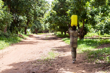 Lonely black African boy in traditional clothing walking on a dirt road with a heavy bright yellow water canister on his head - Powered by Adobe