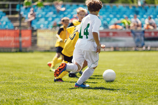 Kids playing football in sunny day. School boys running fast on grass soccer pitch. Junior football players compete in tournament match. Football spectators, stadium crowd in blurred background